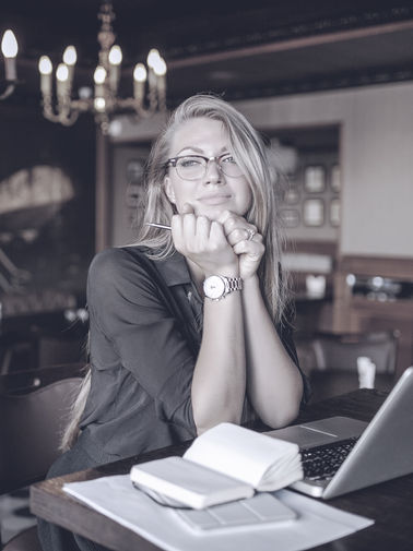 Woman in glasses sits at cafe table, pensively holding pen, laptop and notepad nearby.