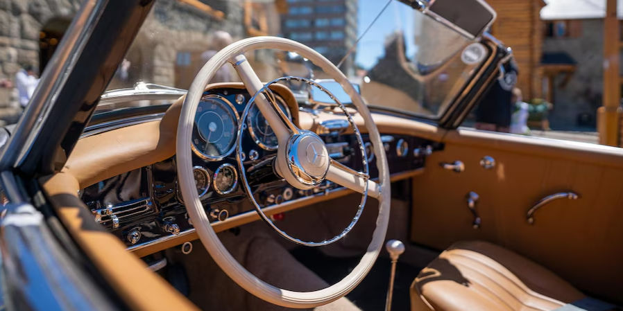 Vintage car interior with tan leather, chrome details, and a classic steering wheel. Background shows blurred stone and wooden buildings.