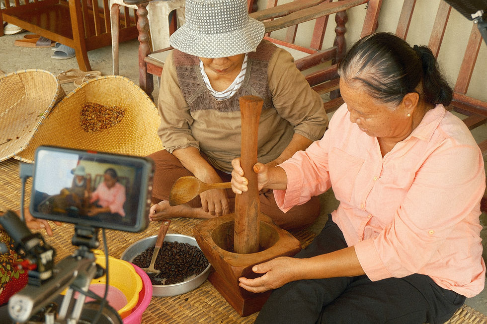 A camera records two women preparing coffee with a wooden mortar beside woven trays.