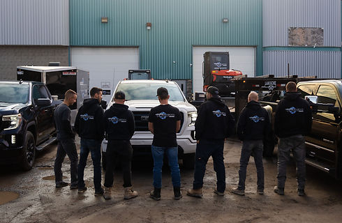 Équipe d'Aménagement Frakar de dos, mettant en valeur le logo sur les chandails, avec un camion, une pelle mécanique et une remorque fermée