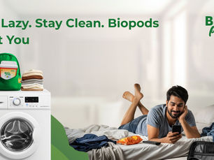 Man lying on a bed, smiling at his phone. Laundry and snacks nearby. Washing machine with detergent in foreground. Text: Be Lazy. Stay Clean.