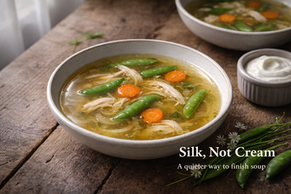 A rustic bowl of clear golden chicken soup with shredded chicken, snap peas, and carrot coins sits on a weathered wooden table, softly lit from the left. The broth has a subtle sheen, with a second bowl and a small ramekin of sour cream blurred in the background. Text overlay reads “Silk, Not Cream” with the subtitle “A quieter way to finish soup.”