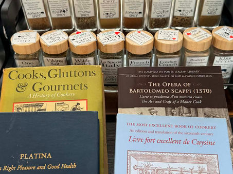 Historic culinary books including Platina and Scappi displayed behind jars of Oak City Spice Blends, representing the connection between Renaissance cooking manuscripts and modern spice craft.
