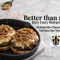 Two cheeseburgers in a cast iron skillet topped with melted seasoned cheese and caramelized onions, with a spoon sprinkling seasoned shredded cheese over the burgers on a marble surface