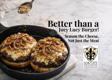 Two cheeseburgers in a cast iron skillet topped with melted seasoned cheese and caramelized onions, with a spoon sprinkling seasoned shredded cheese over the burgers on a marble surface