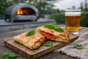 Golden brown homemade calzone resting on a rustic wooden board with fresh basil leaves and a glass of beer, photographed outdoors with a blurred backyard pizza oven in the background.