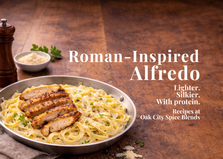 Roman-inspired chicken fettuccine Alfredo in a stainless steel skillet, topped with sliced herb-seasoned chicken, freshly grated Parmesan, and parsley, set on a rustic wooden table with a tall pepper grinder and space for text on the right.