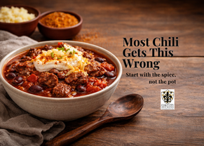 Bowl of steak and two-bean chili topped with sour cream and shredded cheese on a wooden table, with seasoning and ingredients in the background