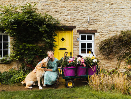 A woman and dog in front of a yellow front door with buckets of dahlias