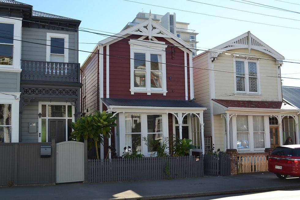 Row of three Victorian houses in urban setting; red and beige homes flank a gray one with ornate balconies and fences, a parked red car.