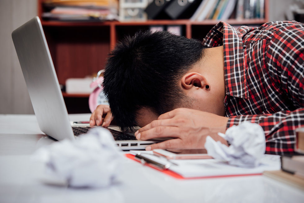 Man in plaid shirt rests head on laptop, surrounded by crumpled papers. Books on shelf, indicating stress or fatigue in office.