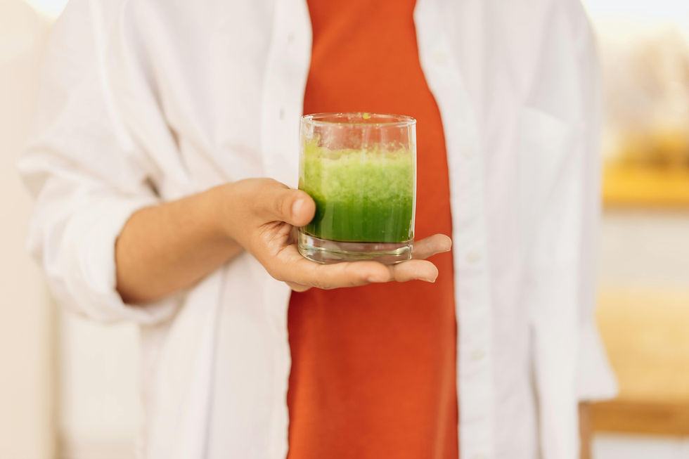 Person in a white shirt and red top holds a glass of green juice. Soft background with warm tones, conveying a fresh, inviting mood.