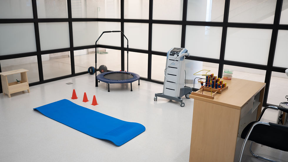 A therapy room with a blue mat, three orange cones, a trampoline, and a medical device on wheels. Wooden toy and table are visible.