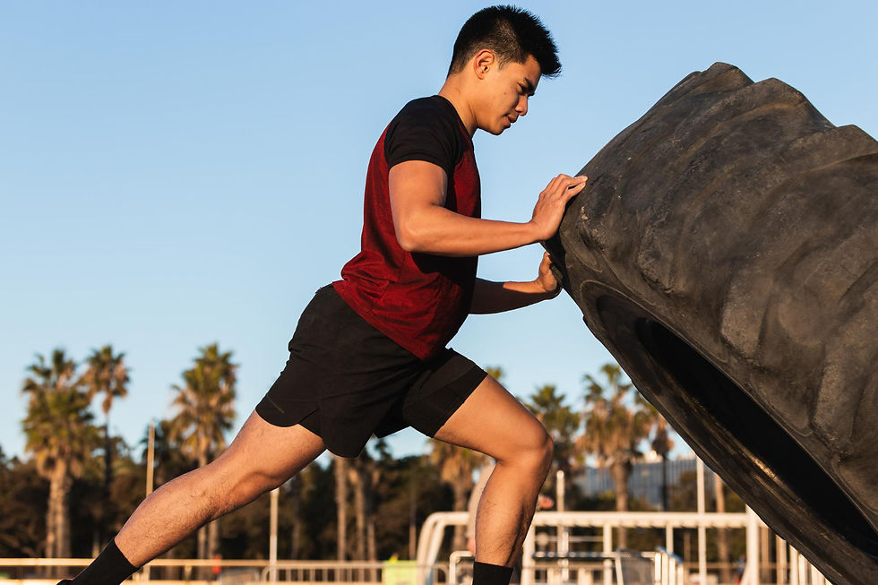 Man in red shirt flips large tire outdoors, focused and determined. Clear blue sky and palm trees in the background.