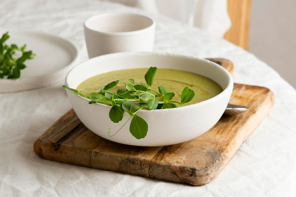 Bowl of green soup with sprigs on top on a wooden board. Nearby is a white cup and a plate. Soft, natural setting.