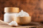 Wooden bowl and spoon with white coconut oil on a rustic wooden surface. A jar is in the blurred background. Warm, earthy tones.