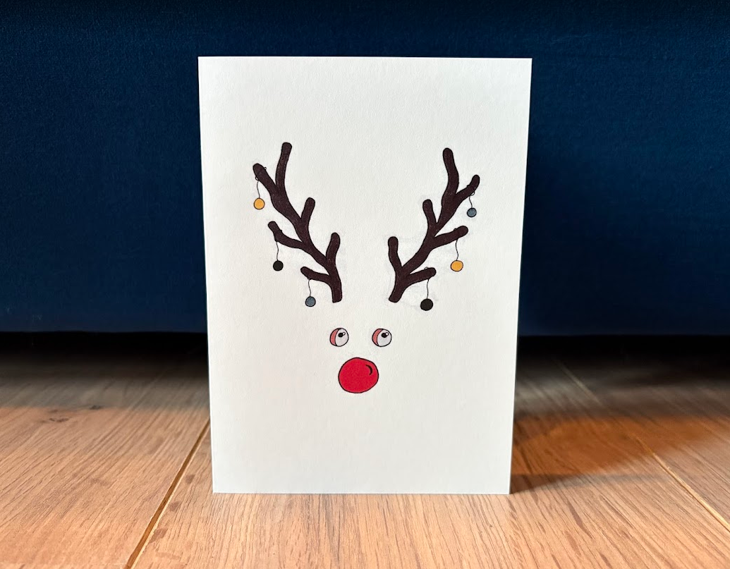 Voorkant wenskaart. Een rendier met gekleurde kerstballen in zijn gewei en een grote rode neus.