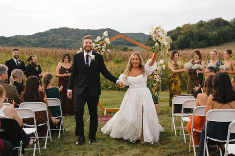 Bride and groom walk smiling, hand in hand down a grassy aisle, as the bride lifts her bouquet into the air.