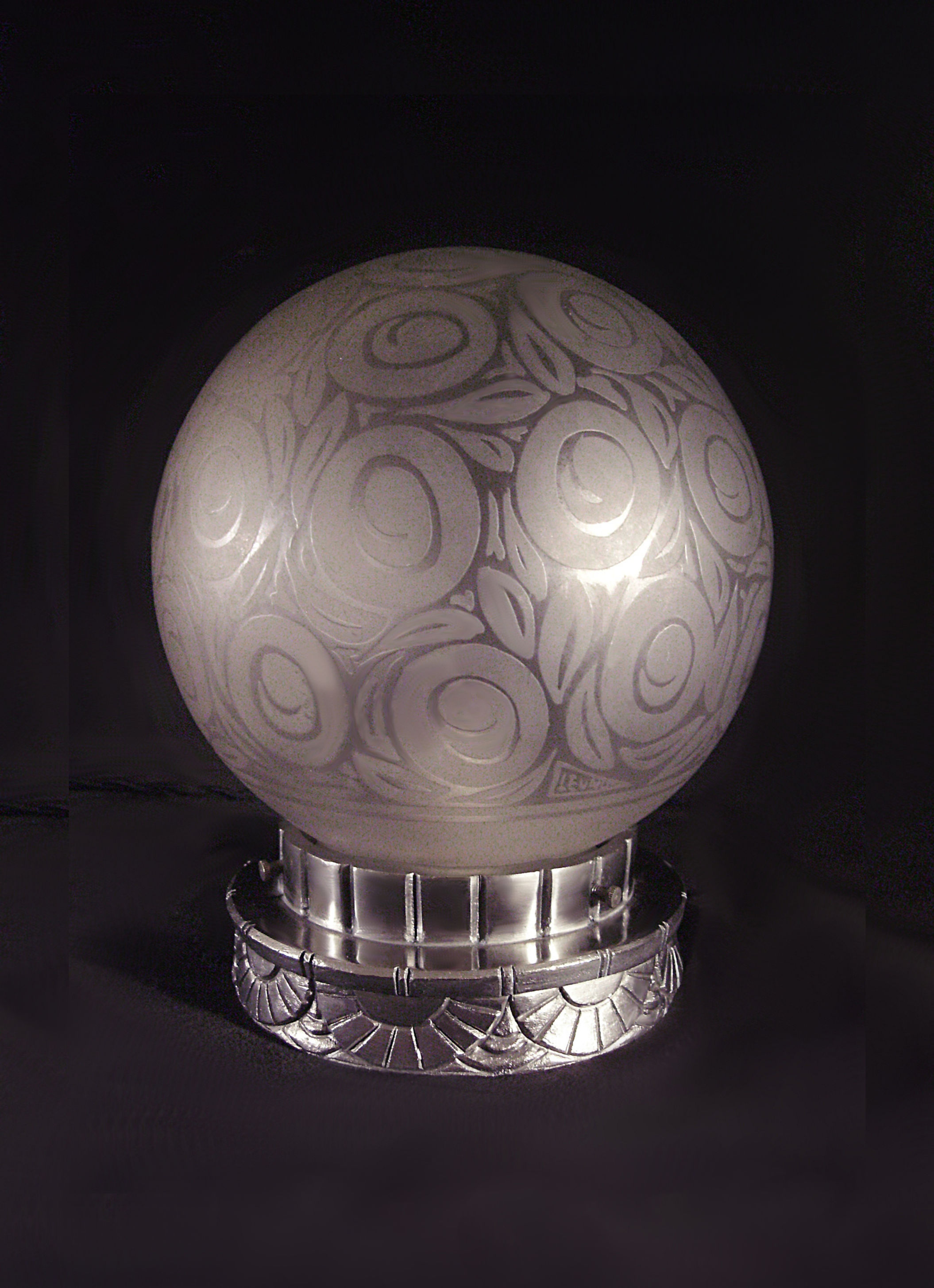 Veilleuse Art déco Leunor en bronze nickelé et globe en verre gravé à décor géométrique de rosaces stylisées, présentée allumée sur fond noir.
