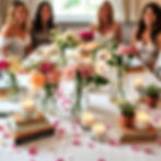 Four women smiling at a table with pink roses, candles, books, and petals. Bright, cheerful mood with a decorated white tablecloth.