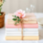 Stack of pastel books tied with twine, topped with a pink flower. Soft background with a wicker basket; elegant and serene setting.