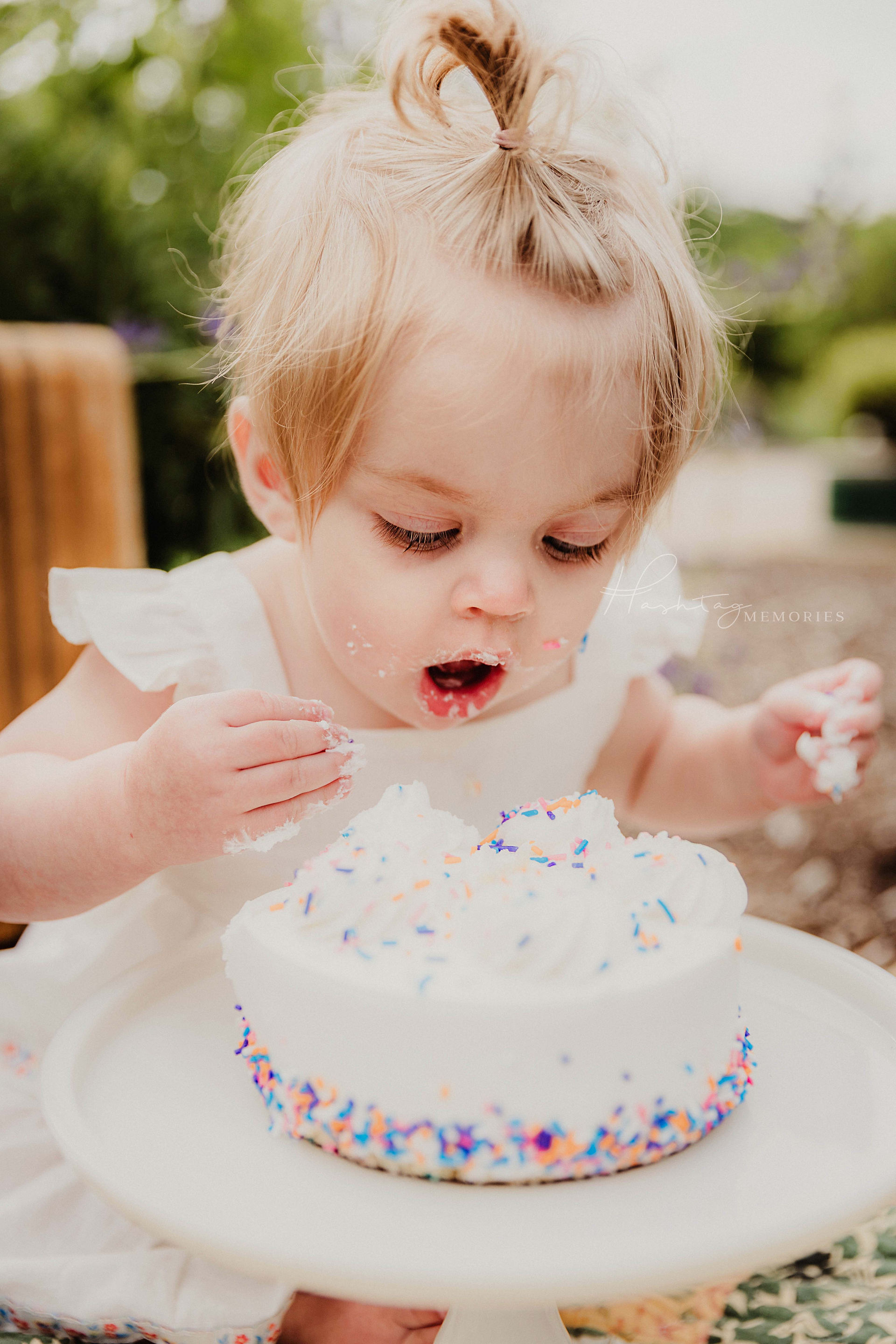 baby girl leans forward to bite into sprinkle birthday cake for her first birthday family photos