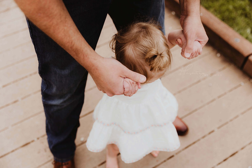 Adult guiding a toddler by the hands on a wooden deck. The child wears a white dress; the adult is in jeans. Mood is caring and tender.