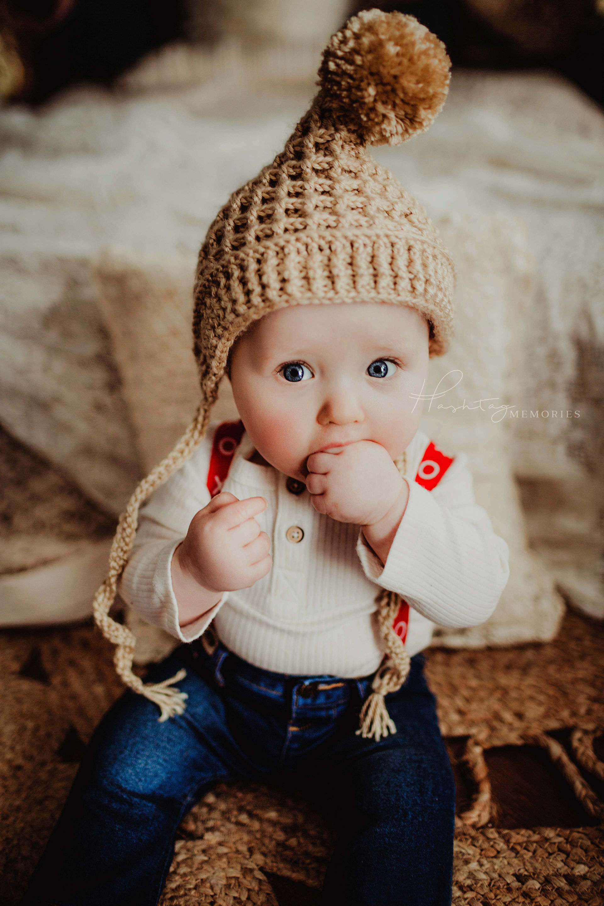 baby boy sits, sucking on his fingers with cute winter hat on, jeans and red suspenders