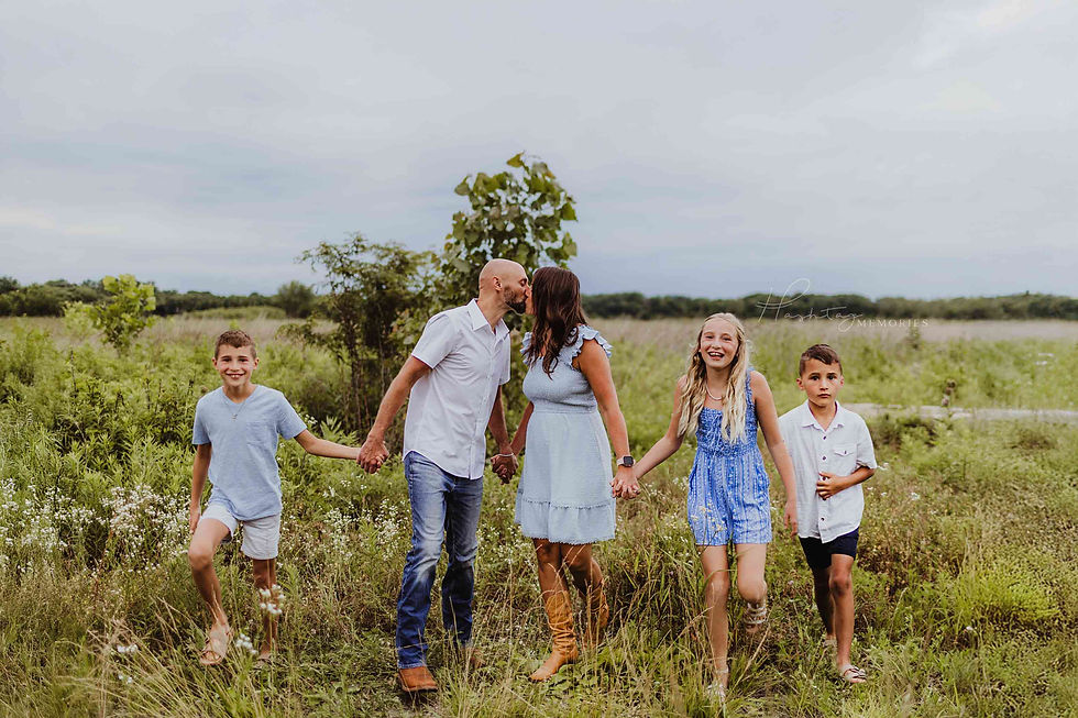 Family of five in a grassy field; parents kiss while holding hands with three smiling children. Hashtag Memories watermark.