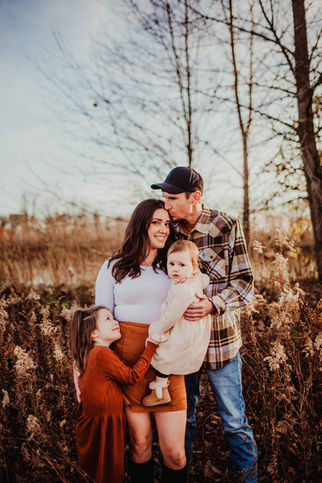 mom holds baby girl in her arms while being kissed by dad and hugged by her older daughter in fall fieds