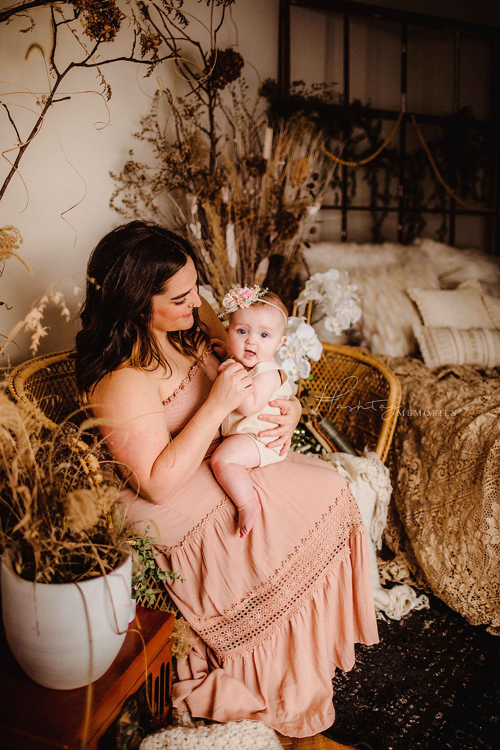 mother holds young baby girl in her arms, wearing boho pink maxi dress and flower headband while sitting on vintage whicker couch with lace