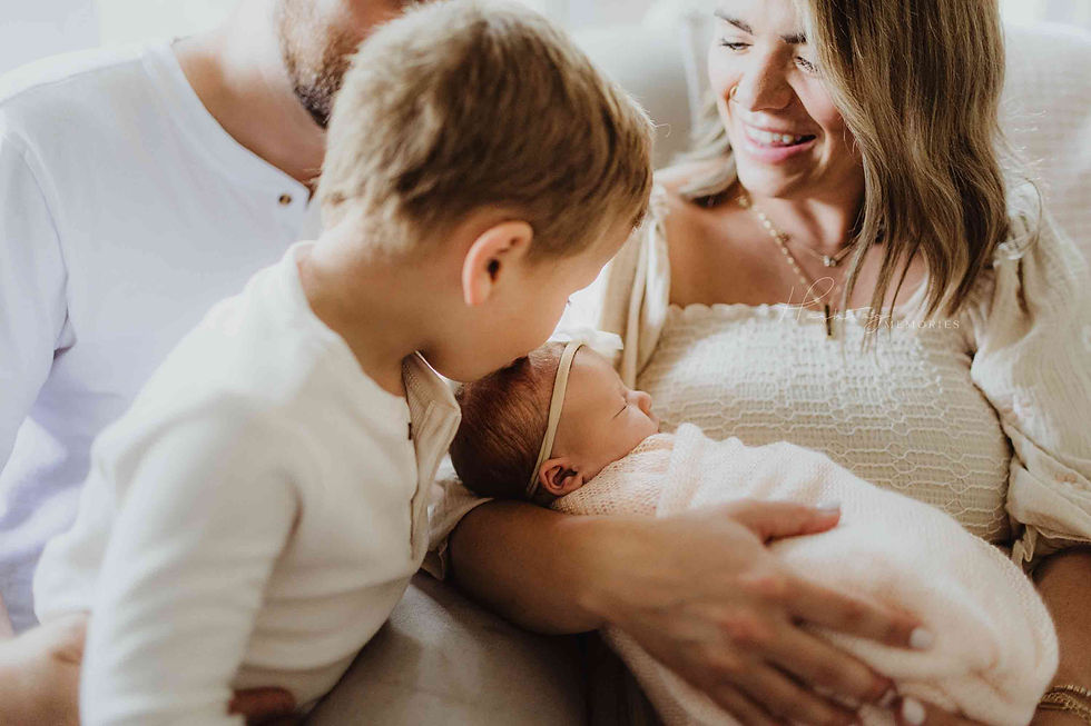 Smiling mother holds a newborn while a young boy leans in and a bearded father sits nearby on a soft couch.