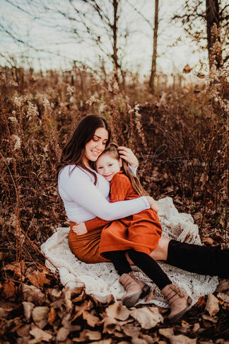 mother wearing white and burnt orange miniskirt sits in fall fields holding young daughter in her lap