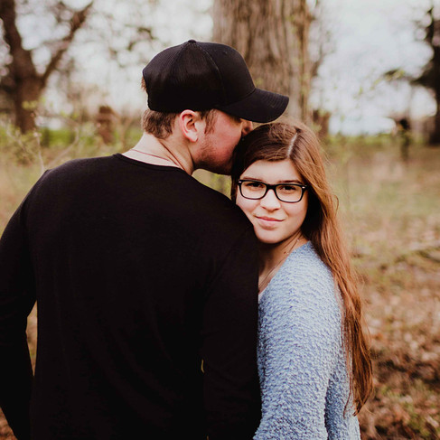 engaged couple stands together in carmel indiana woods. They are holding hands, she is facing the camera with a smile, his back is to the camera, head turned to kiss the top of her head.