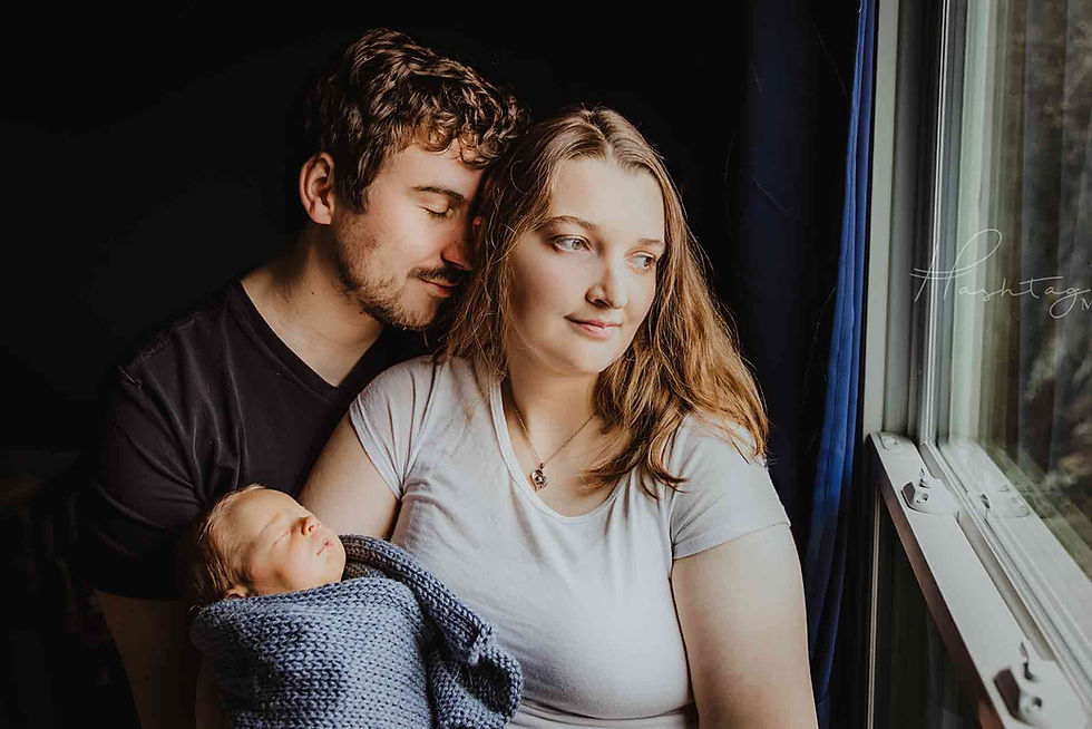 A couple cuddles their newborn wrapped in a blue blanket by a window. The setting is serene with soft natural light.