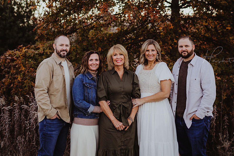 A family of five stands smiling in front of autumn foliage. They wear casual fall clothing in earthy tones. The mood is warm and joyful.