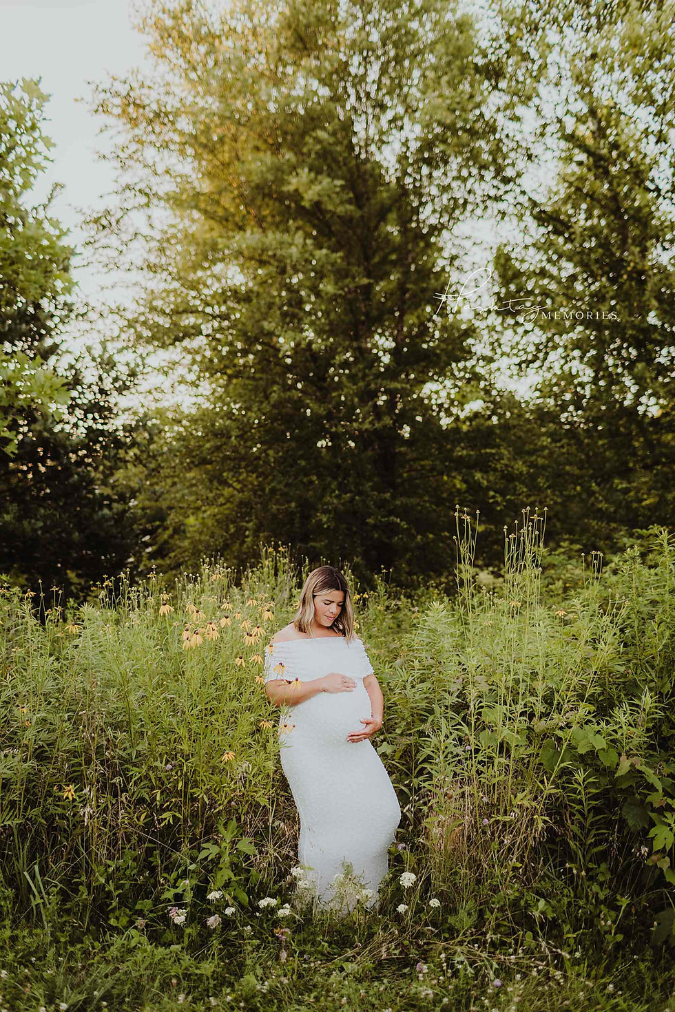 Pregnant woman in a white dress stands in a wildflower meadow, hands on belly, with tall trees behind and a Memories watermark.