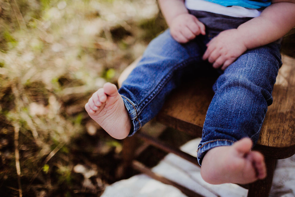 Toddler in blue jeans sits on a wooden bench outdoors, bare feet dangling against a blurred green background.