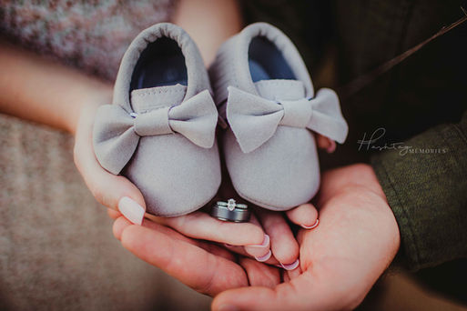 women's hands hold baby shoes and stacked his/her wedding bands ontop of a man's hands announcing they are expecting a baby
