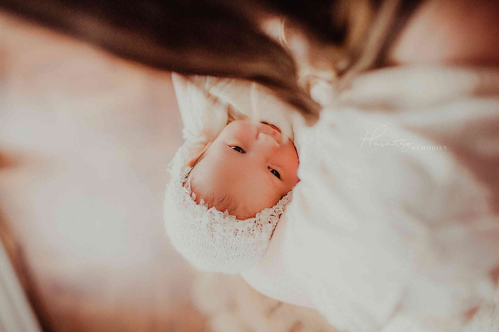 A baby, wrapped in white, looks at the camera with a calm expression. Soft lighting and blurred background create a serene mood.