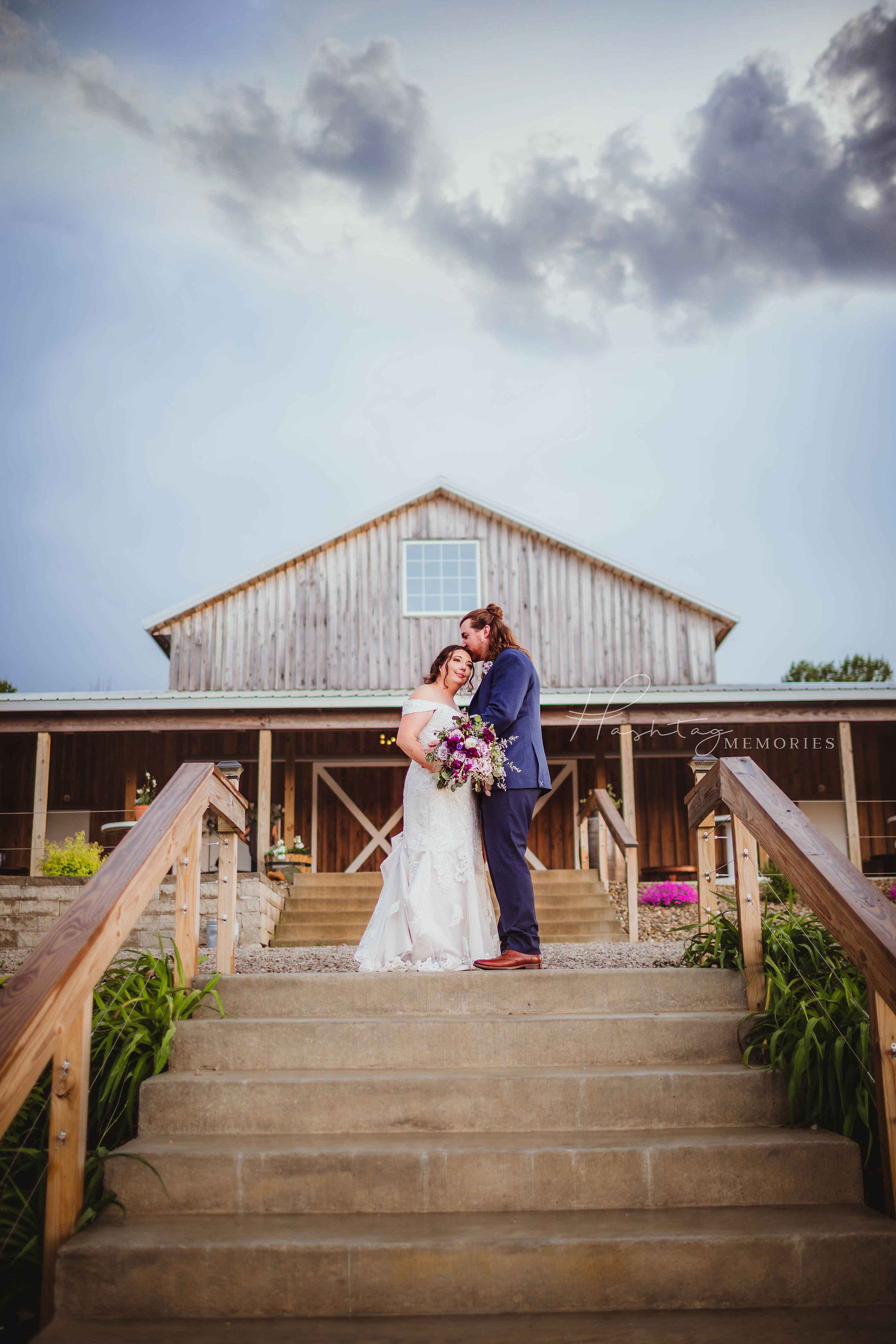 The Barn at Hamner Ridge Edinburgh, Indiana wedding venue. Bride and groom stand in front of the wedding barn with large porch and bride and groom kissing in the middle on top of the entry stairs