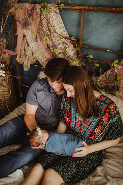 mother and father hold newborn baby girl in their arms together, sitting on a bed. Baby wrapped in a light blue accents and headband