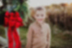 Smiling child in a brown sweater stands by festive greenery with a red bow. Snow gently falls in a grassy outdoor setting.