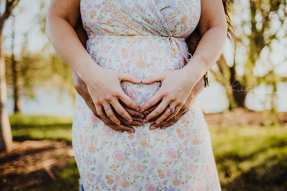Couple announces their pregancy by making a heart with their hands on the mother's belly. Father is standing behind the mother. Both sets of hands for a heart and both wedding bands can be seen as well. Carmel, Indiana Maternity Photo by Midwest Photographer