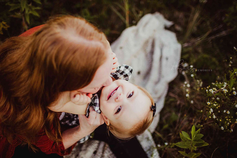 A mother embraces her smiling child on a picnic blanket among greenery. The child wears a black and white checkered outfit.
