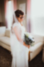 bride stands in bridal suite in front of vintage couch wearing a white wedidng dress with sleeves and sparkle veil holding her bridal flowers. Modest wedding dress.