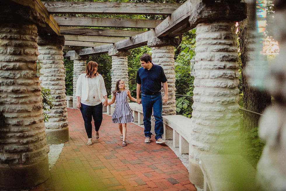 A family walks hand-in-hand on a brick path under a rustic pergola. The setting is lush with greenery, creating a serene mood.