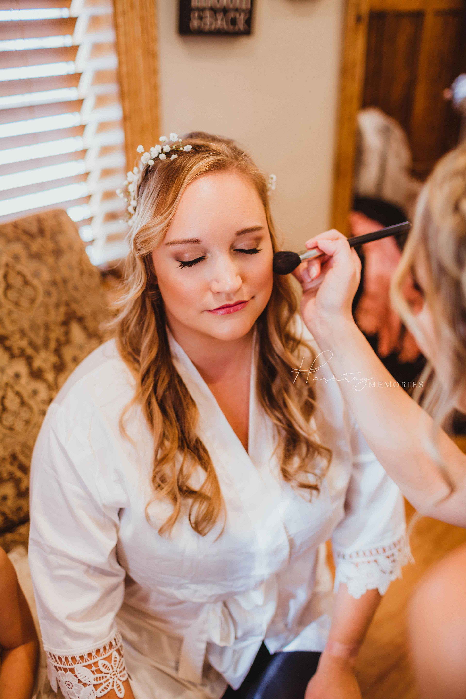 Sylvan Cellars Bride on her wedding day, wearing her hair down with white silk robe in the bridal suite with baby's breathe florals in her hair as her make up is being done