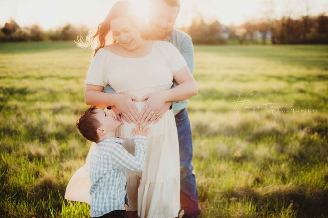mother and father have their hands on mother's pregnant belly while their son looks up at them and touches the belly in a sunrise field in carmel indiana