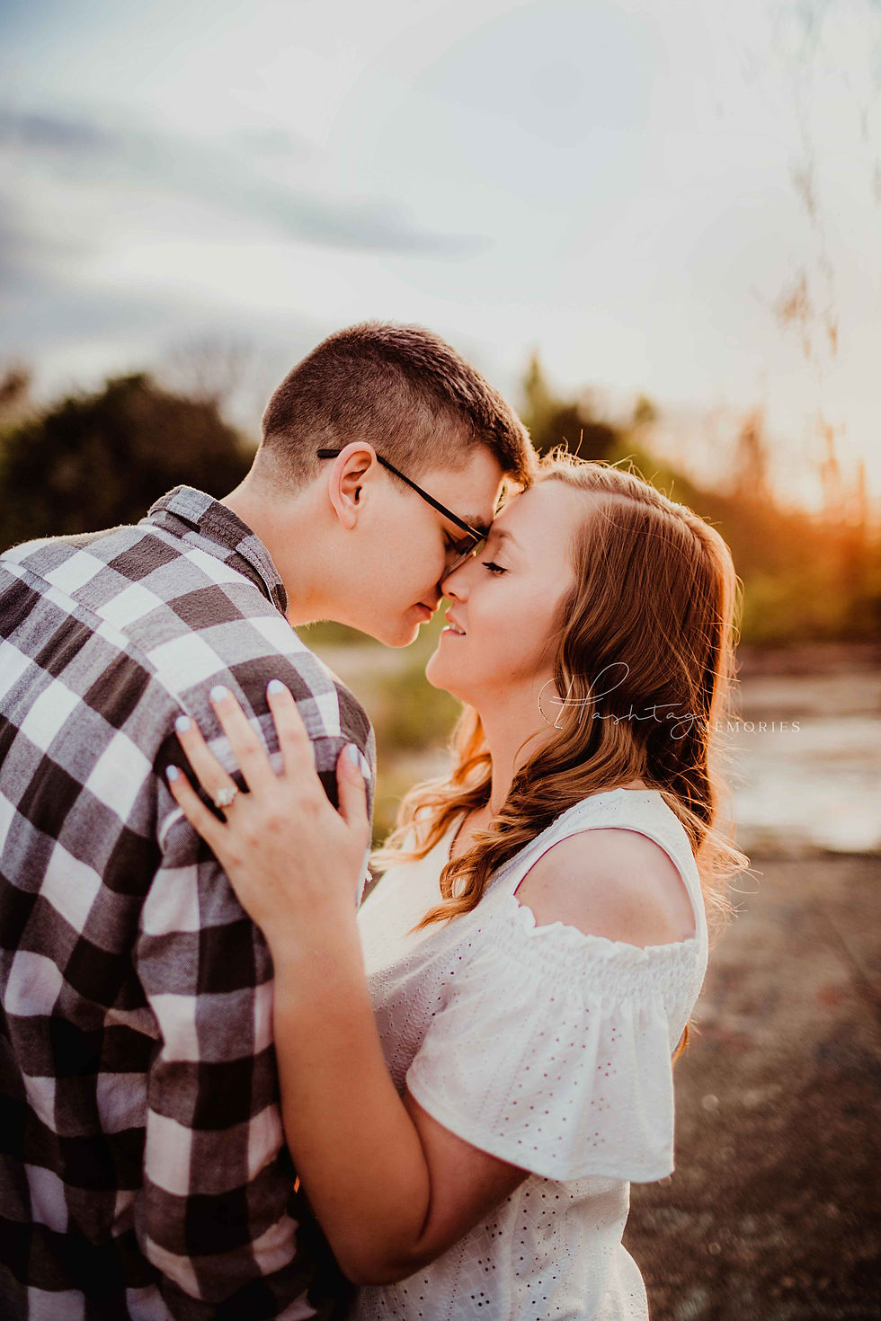 couple kisses in urban jungle setting with bright sunset behind them. Boho vibes as they kiss, she wears a white off the shoulder shirt with jeans and sandles. he wears long sleeve black and white buffalo plaid with 50s style eye glasses